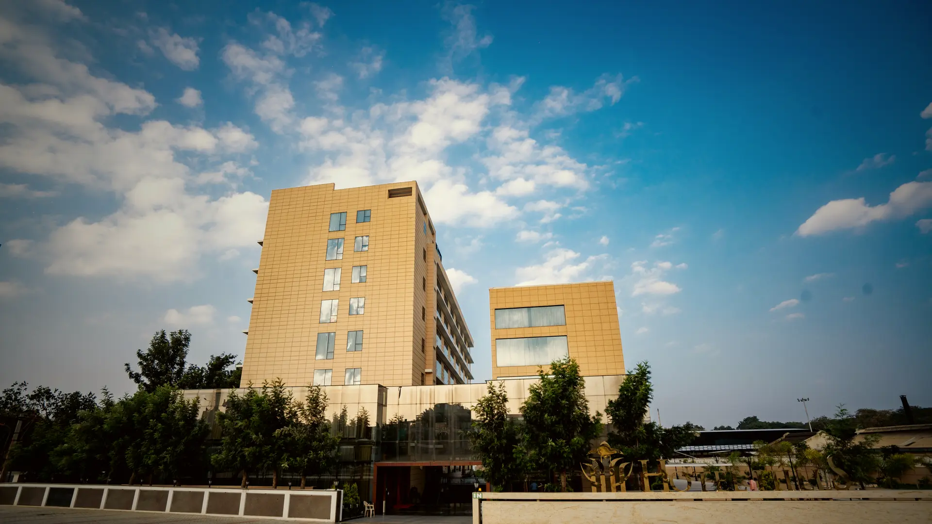 The tower of Hotel Hills Hosur from the forecourt in the late afternoon, the hotel name at its top, the paved forecourt in front