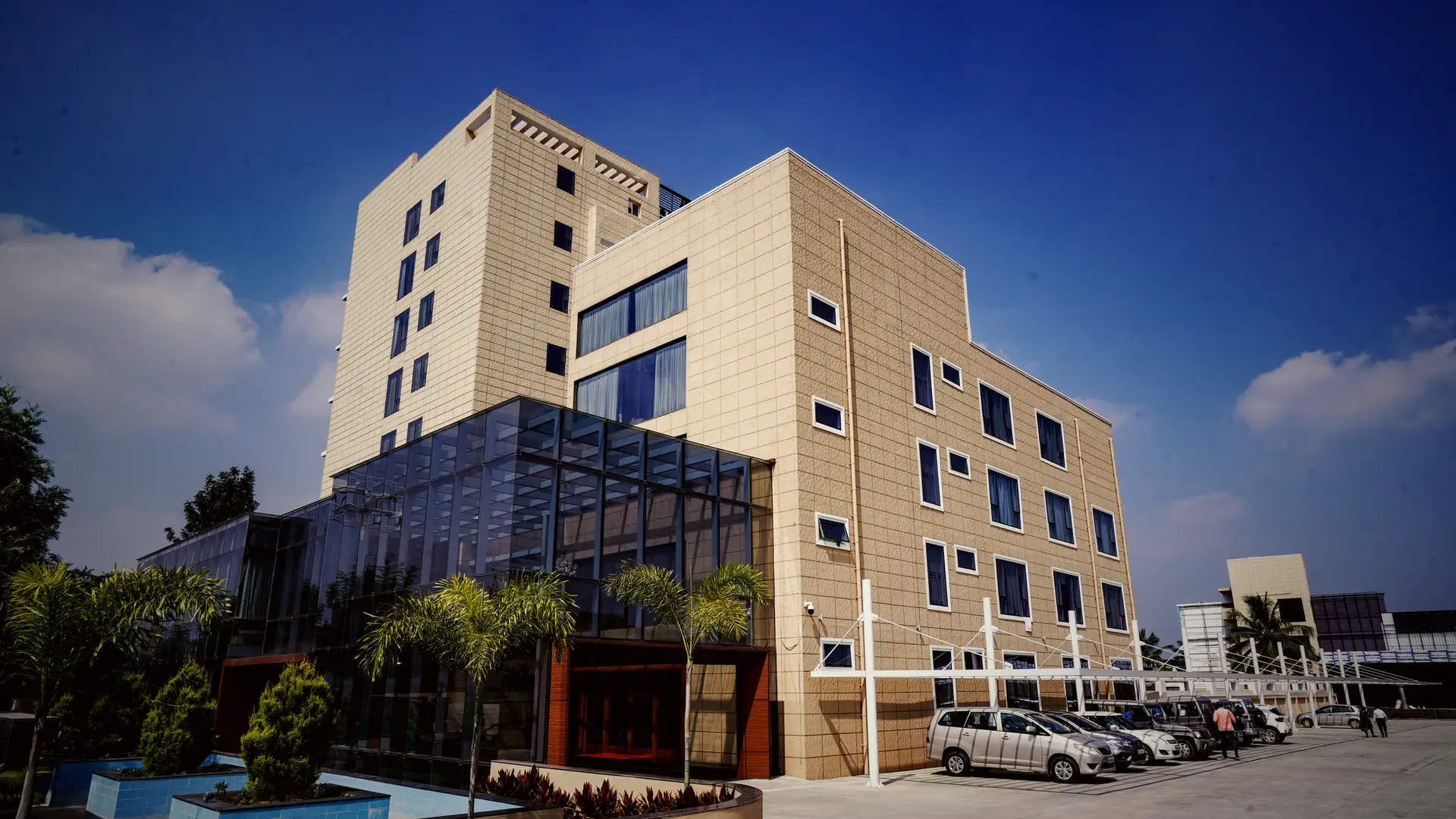 The glazed wing of Hotel Hills Hosur from the forecourt, its sloping glass roof against the stone clad tower, palms and parked cars in front