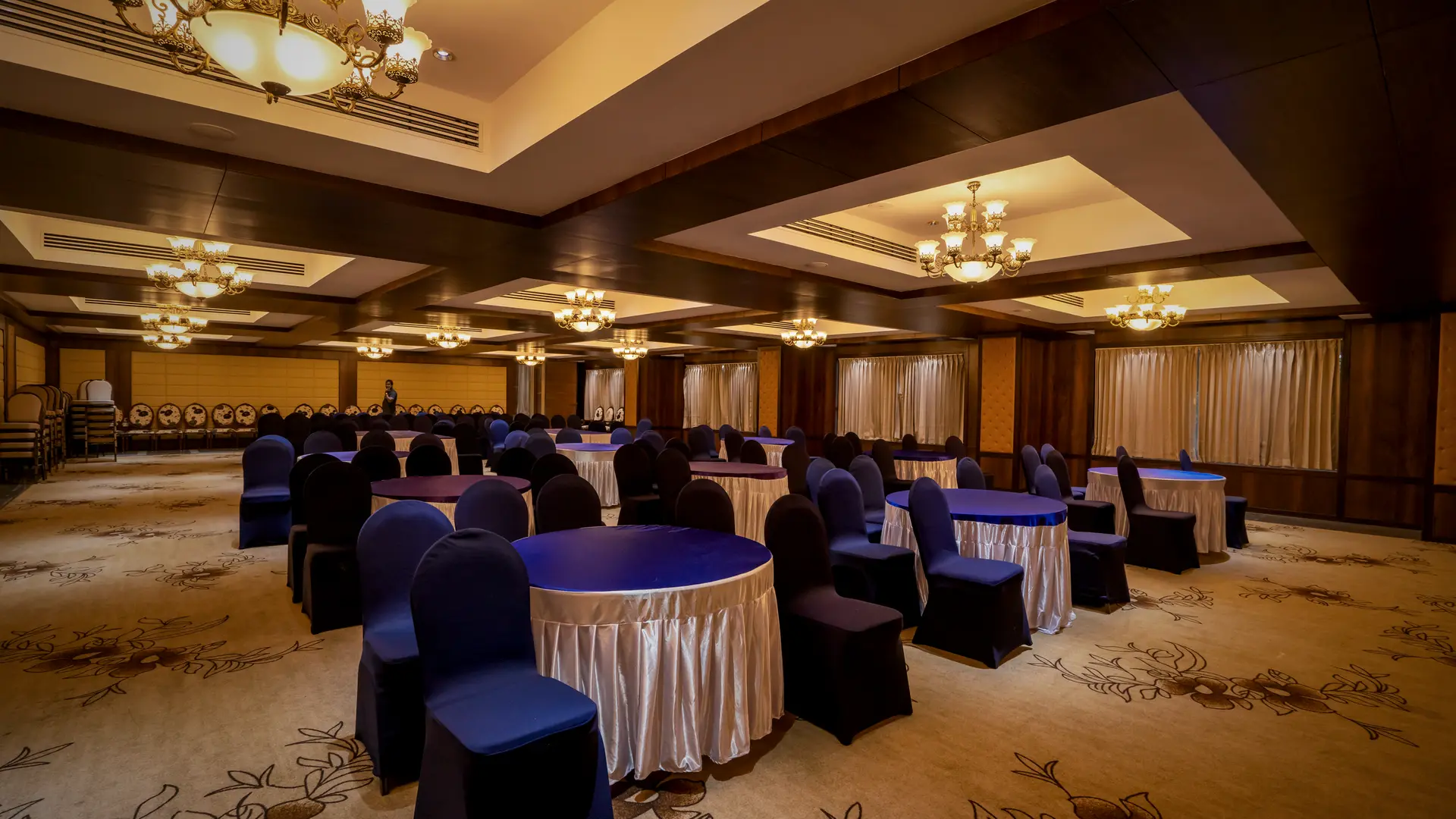 The convention hall at Hotel Hills Hosur set with round tables in blue and cream covers, chandeliers in coffered ceiling panels above