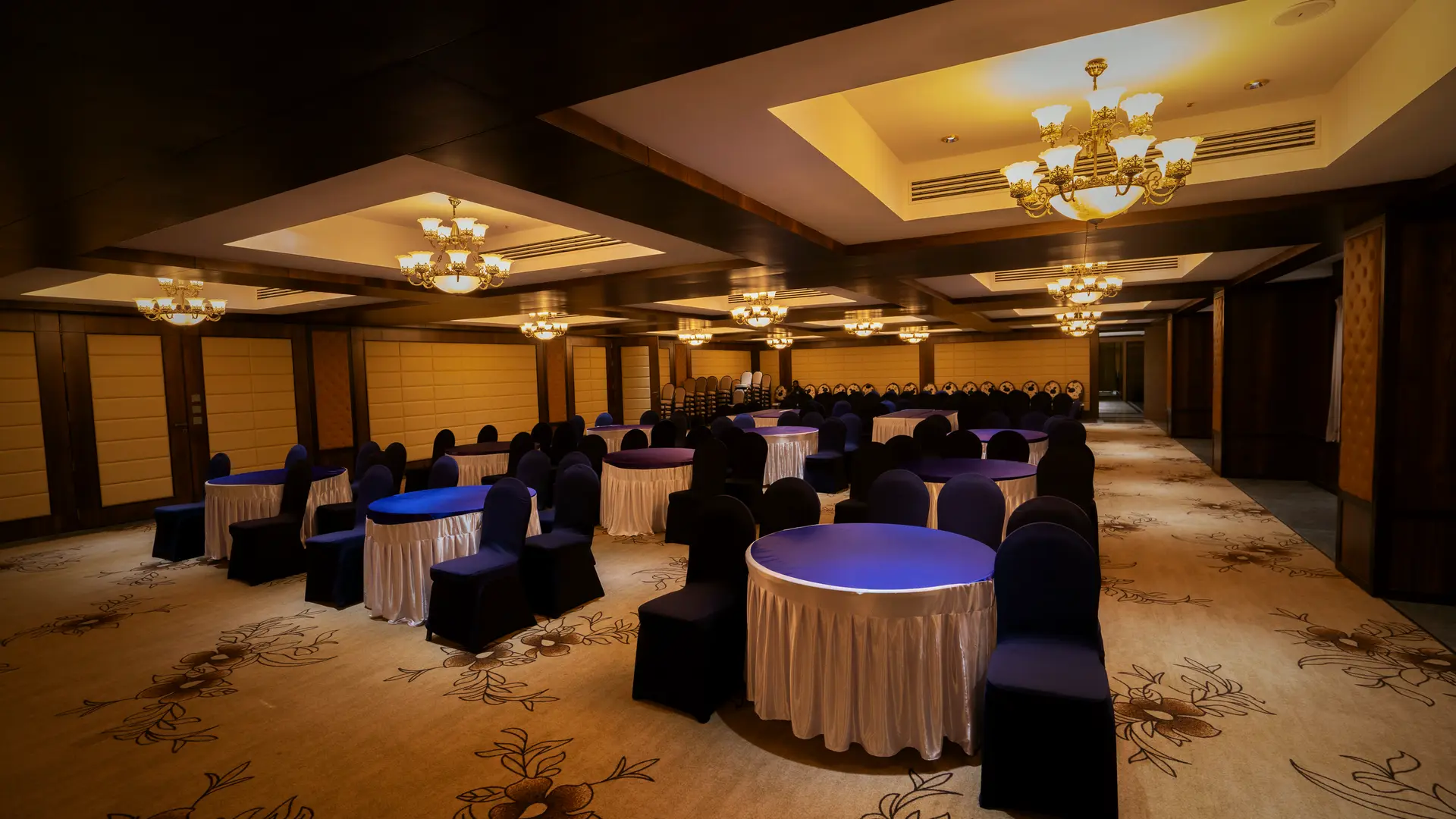 The convention hall at Hotel Hills Hosur from the side wall, round tables with blue chair covers running the length of the room