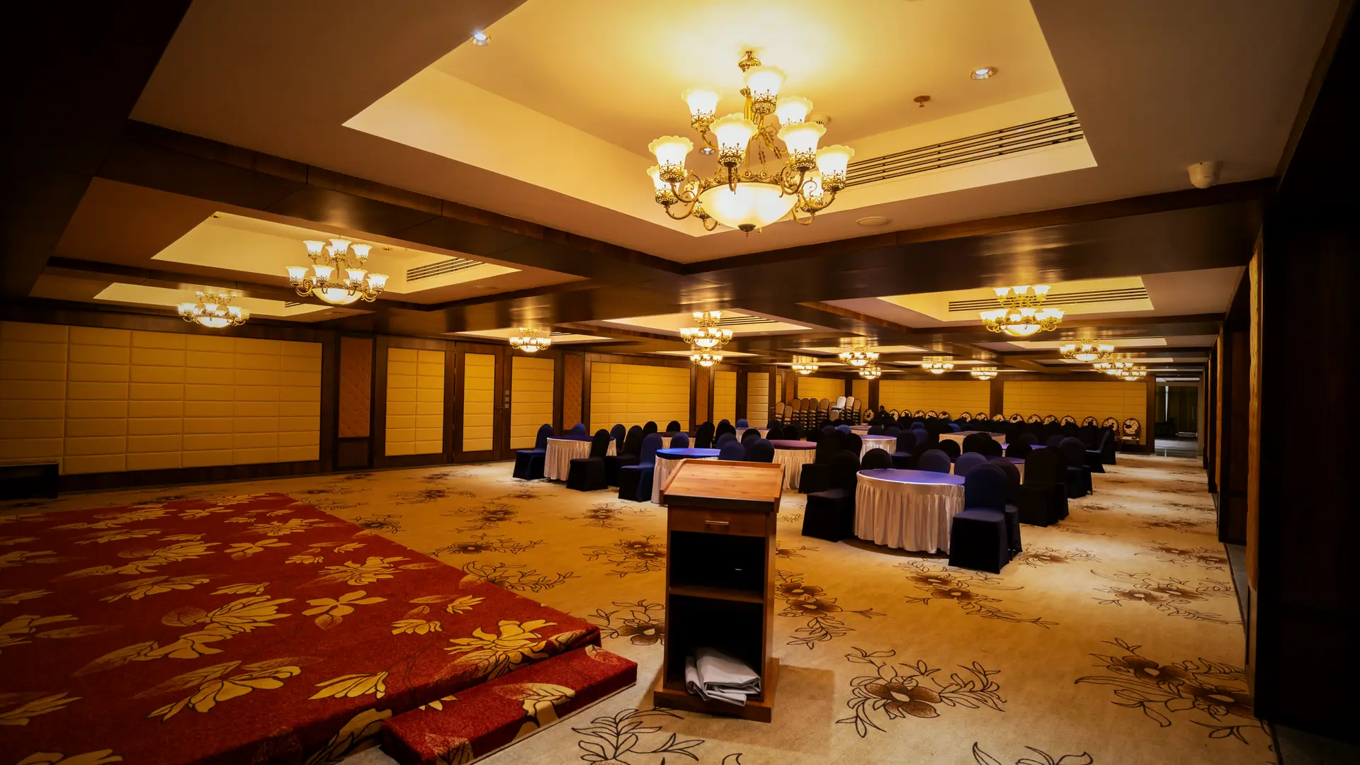 The convention hall at Hotel Hills Hosur from the stage end: a lectern on a red carpet, round tables beyond it, chandeliers above