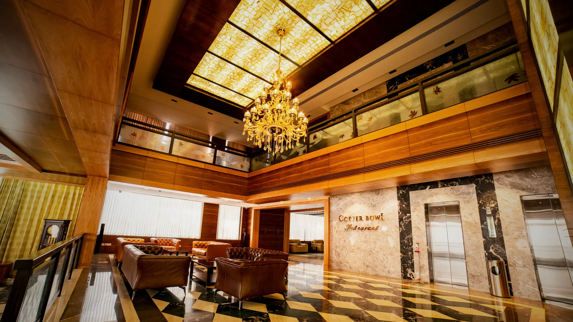 The double height lounge at Hotel Hills Hosur: a chandelier under a lit ceiling panel, a timber gallery above, the marble wall with the Copper Bowl lettering and the lifts beside it