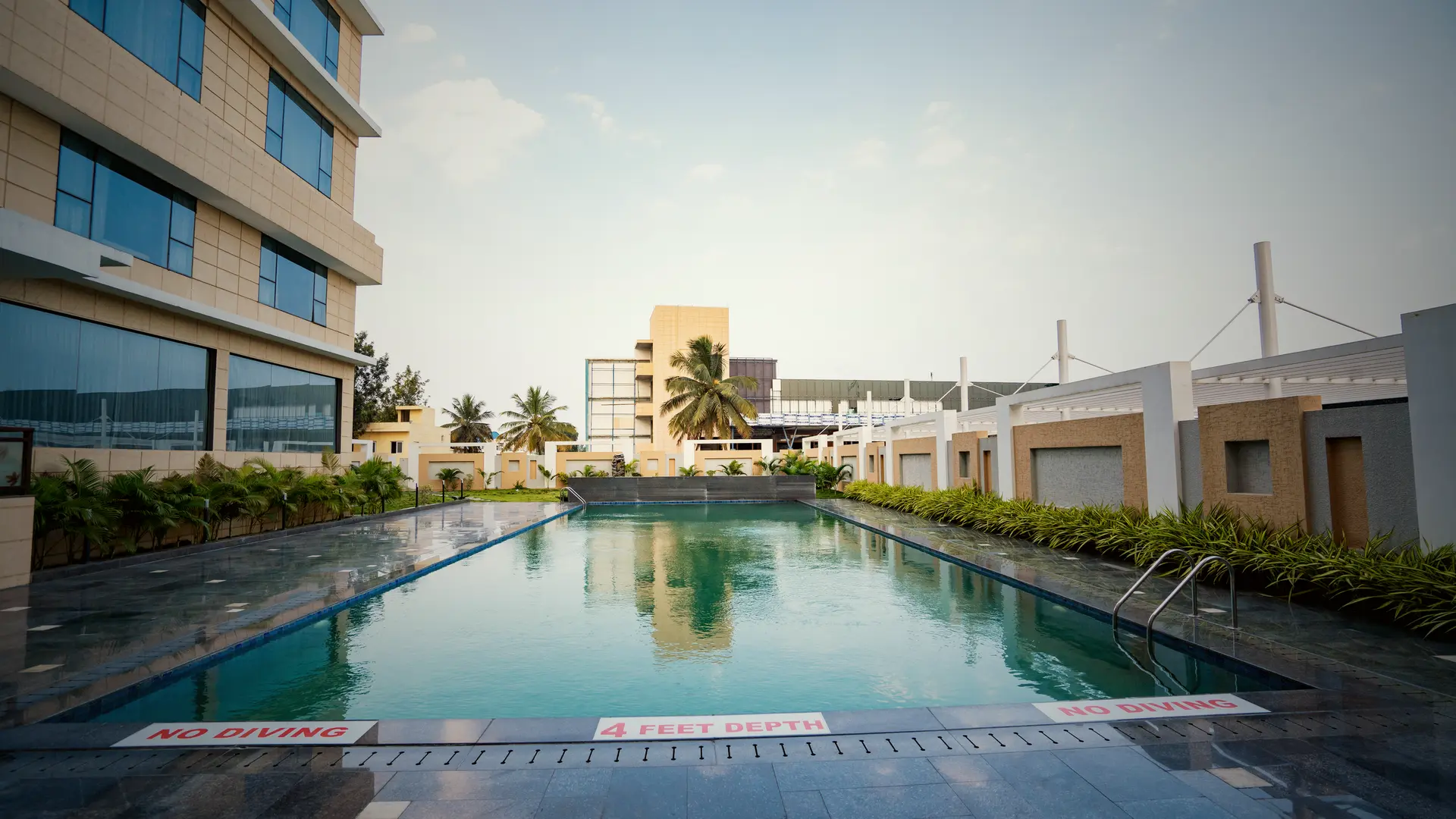 The pool at Hotel Hills Hosur looking down its length, the glass of the tower along the left side and a hedge along the right