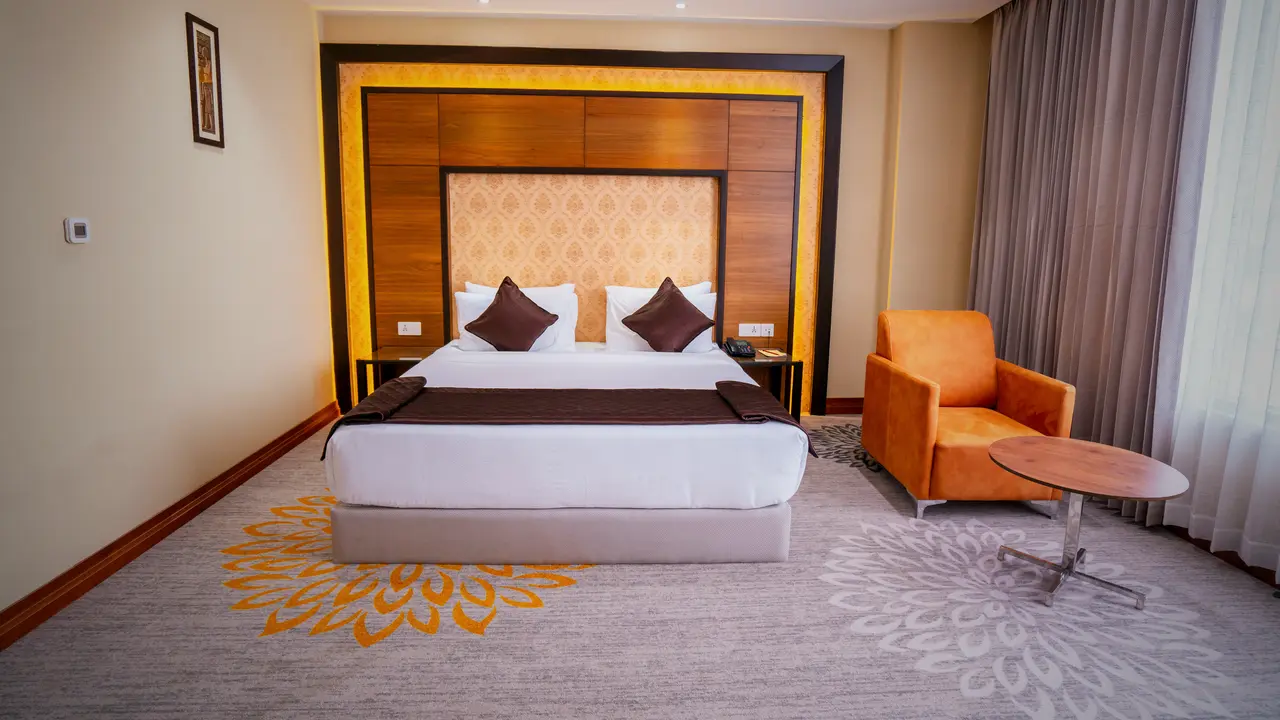 A king room at Hotel Hills Hosur: a king bed against a lit timber headboard panel, a patterned carpet and an orange armchair by the window