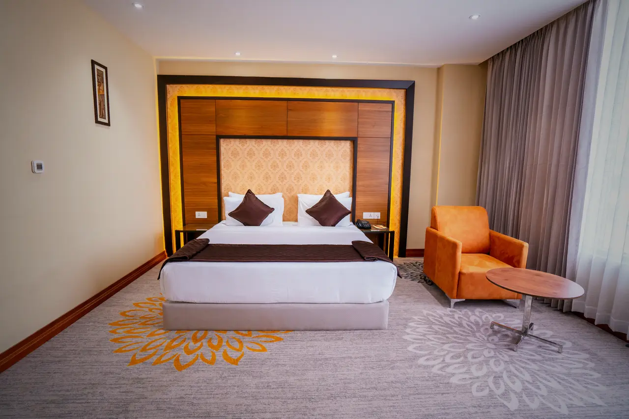 A king room at Hotel Hills Hosur: a king bed against a lit timber headboard panel, a patterned carpet and an orange armchair by the window