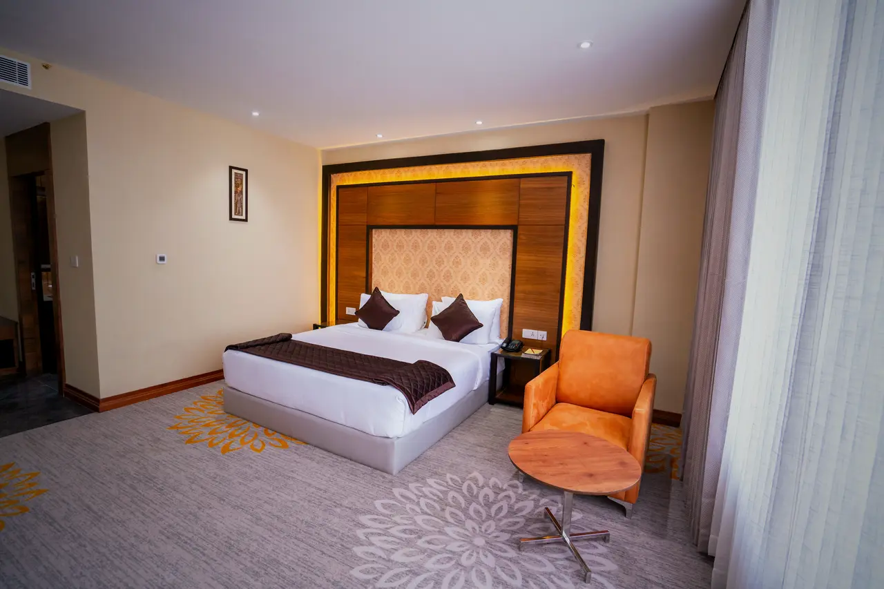 A king room at Hotel Hills Hosur: a king bed against a lit timber headboard panel, a patterned carpet and an orange armchair by the window
