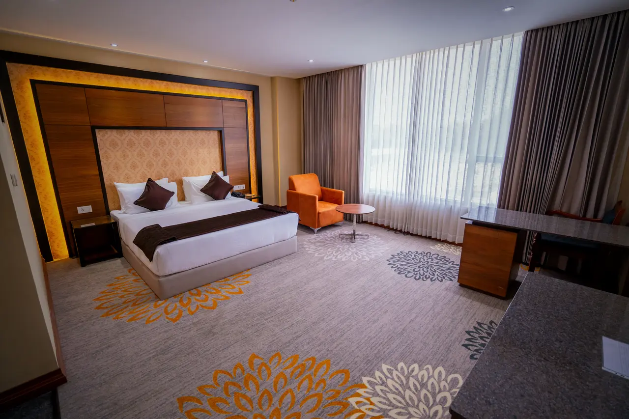 A king room at Hotel Hills Hosur: a king bed against a lit timber headboard panel, a patterned carpet and an orange armchair by the window