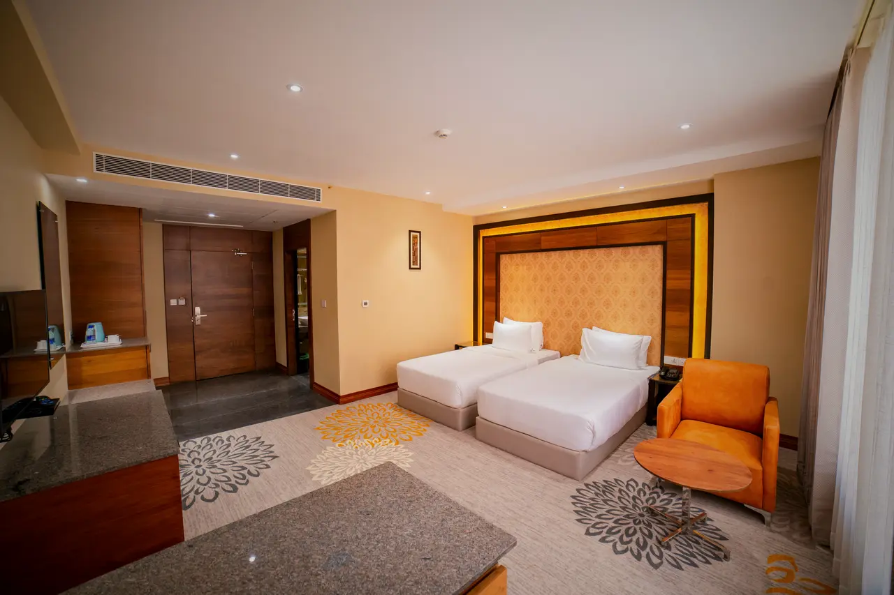A twin room at Hotel Hills Hosur: two single beds against a lit timber headboard panel, an orange armchair, and the entrance door