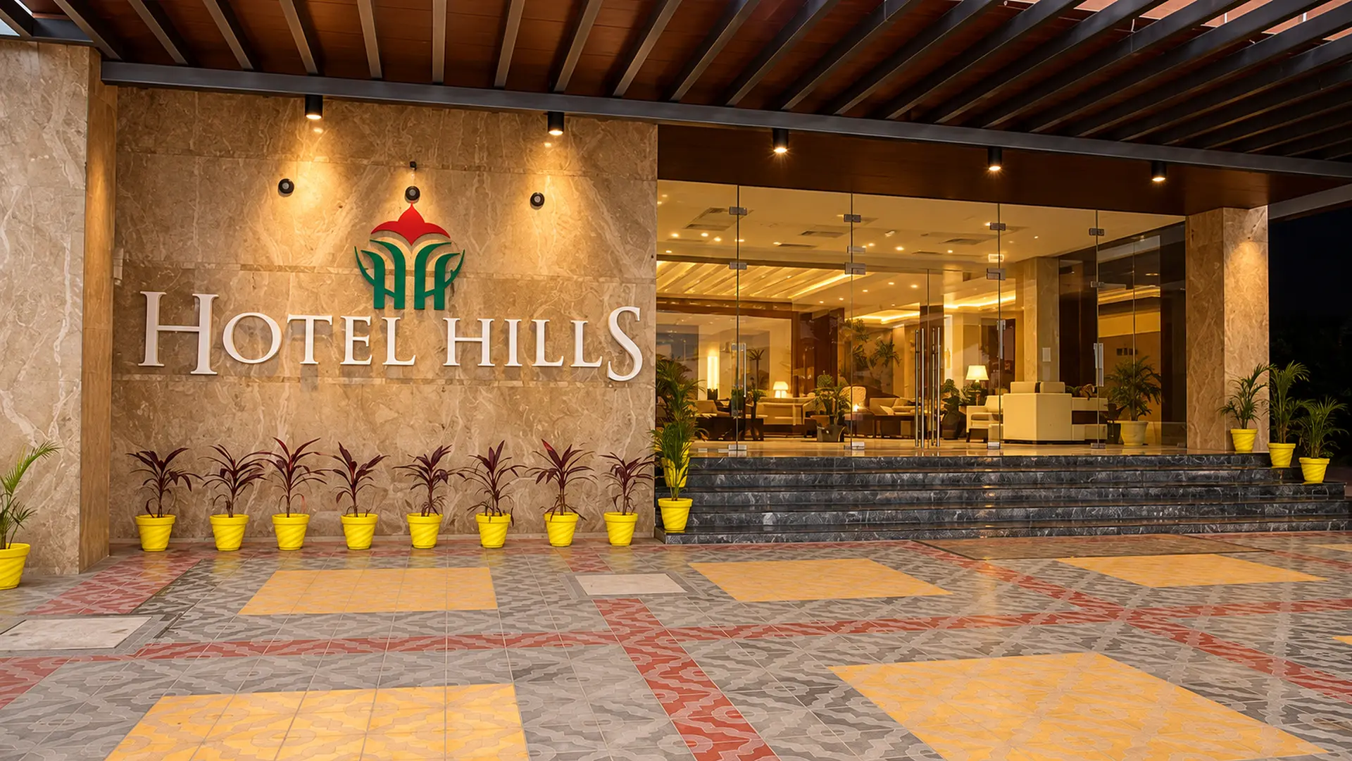 The entrance of Hotel Hills Tirupattur at night: the name in metal letters lit on a stone wall, under a slatted timber pergola, with planters along the front