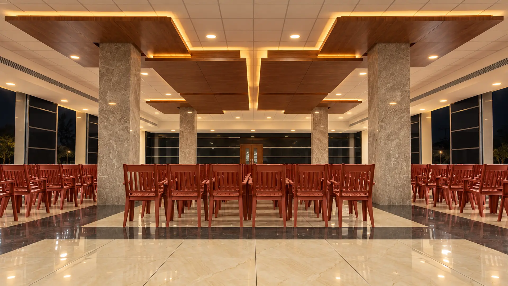 The banquet hall at Hotel Hills Tirupattur: rows of dark red wooden chairs facing the far end, marble columns either side, and a panelled timber ceiling
