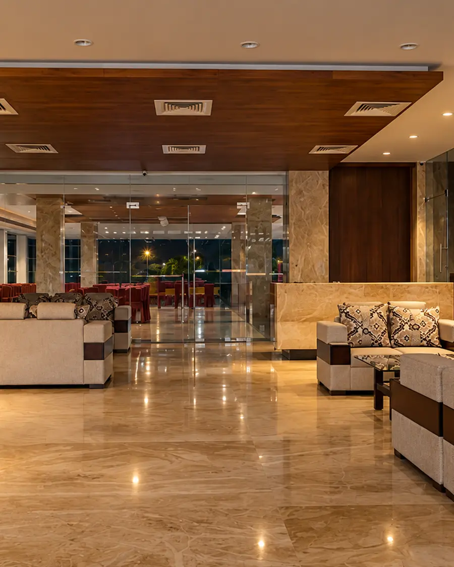 The lobby at Hotel Hills Tirupattur at night: low sofas either side of a polished stone floor, marble columns, and the glass front dark behind them