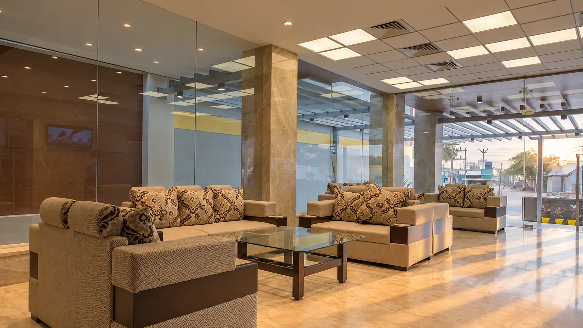 The lobby at Hotel Hills Tirupattur in the morning: daylight through the glass front onto the sofas and the polished stone floor