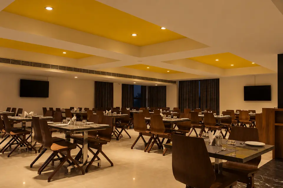The restaurant at Hotel Hills Tirupattur in the evening: tables laid with white cloths and glassware under a yellow ceiling panel