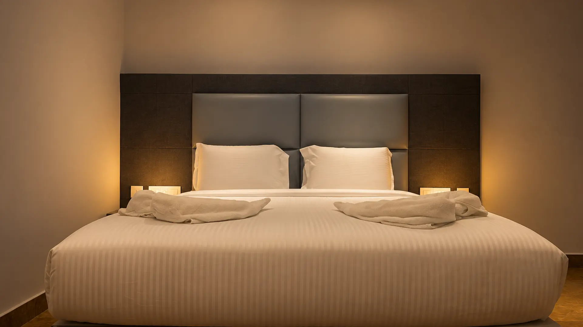 A king bed at Hotel Hills Tirupattur with a grey padded headboard, white bedding and folded towels, against a plain wall