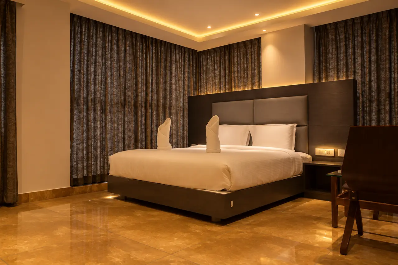 The suite at Hotel Hills Tirupattur: a king bed against a timber headboard, floor length curtains drawn across the window wall, and a writing desk beside it